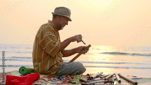 Senior man sits on the beach and making flute by hands, close-up.