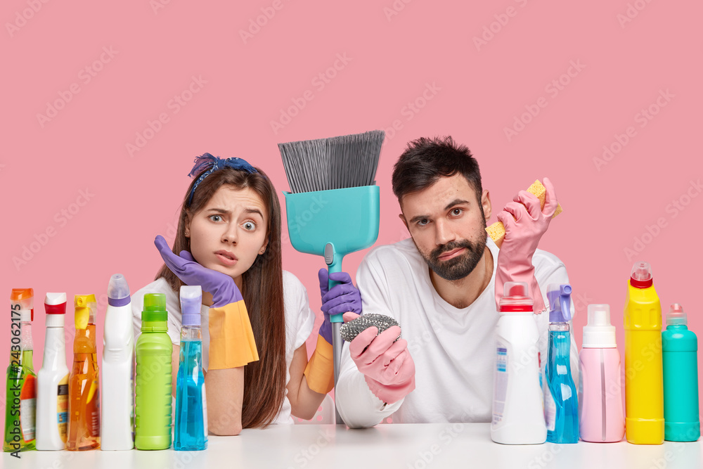 Displeased Caucasian woman and man look with displeasure, feels fatigue after spring cleaning in house, use broom, detergents and sponge for bringing apartment in order, isolated over pink wall Displeased Caucasian woman and man look with displeasure, feels fatigue after spring cleaning in house, use broom, detergents and sponge for bringing apartment in order, isolated over pink wall