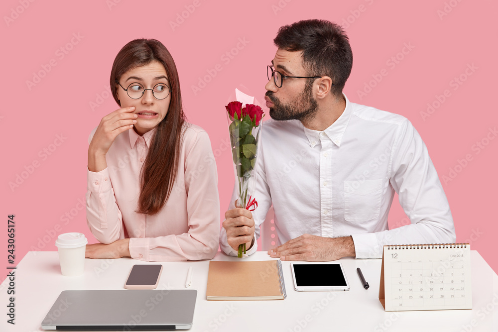 Photo of puzzled attractive woman clenches teeth, looks with shy expression at handsome guy who gives her bouquet of roses, have date at work place, isolated over pink background. Romance concept Photo of puzzled attractive woman clenches teeth, looks with shy expression at handsome guy who gives her bouquet of roses, have date at work place, isolated over pink background. Romance concept