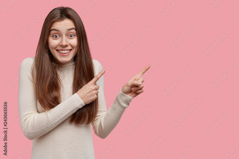 Horizontal shot of pleased joyful European woman shows customers cool product, points aside at blank screen, wears casual clothes, smiles broadly, isolated over pink background shows something awesome Horizontal shot of pleased joyful European woman shows customers cool product, points aside at blank screen, wears casual clothes, smiles broadly, isolated over pink background shows something awesome
