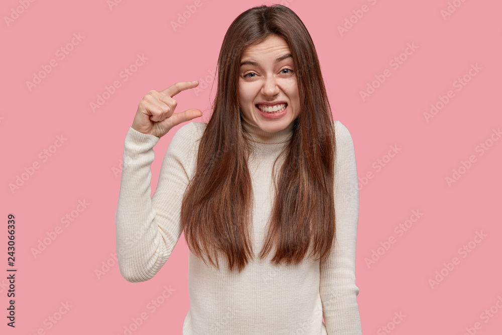 Size matters. Pleased brunette young woman demonstrates very tiny object, smiles positively, wears casual white sweater, poses against pink background, shapes small thing. Body language concept Size matters. Pleased brunette young woman demonstrates very tiny object, smiles positively, wears casual white sweater, poses against pink background, shapes small thing. Body language concept