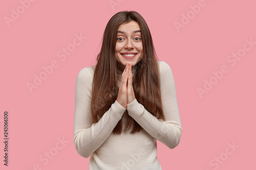 Pleased smiling lady has pleading expression, keeps palms pressed together, prays for everything good, has long dark hair, wears casual clothes, asks for help, isolated over pink background.