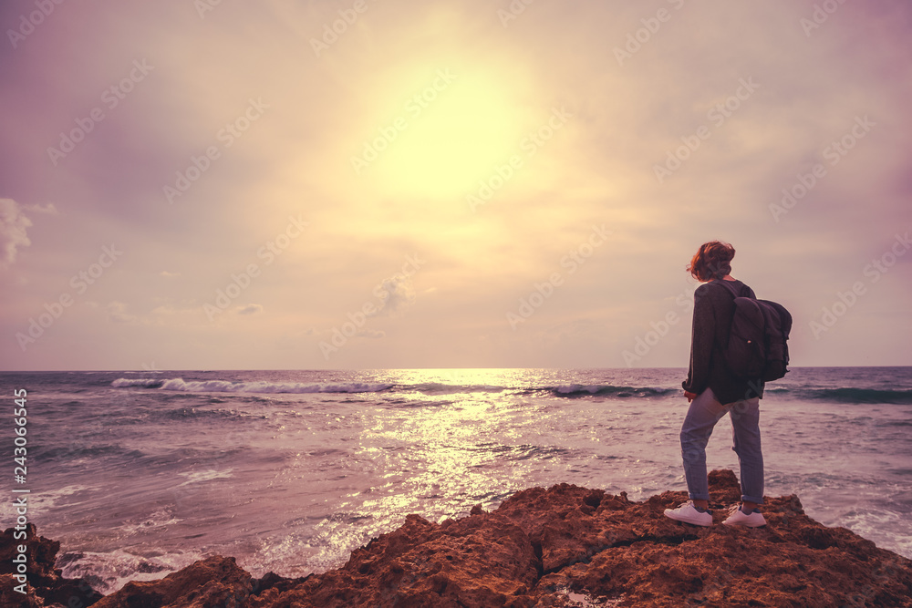 Fototapeta premium A girl with a backpack is standing with her back on the stormy seashore, an image with retro toning