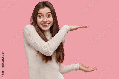 Body language concept. Glad Caucasian woman shows size of box with two hands, smiles gladfully, wears casual white sweater, isolated over pink wall, expresses positive emotions, gestures actively © wayhome.studio 