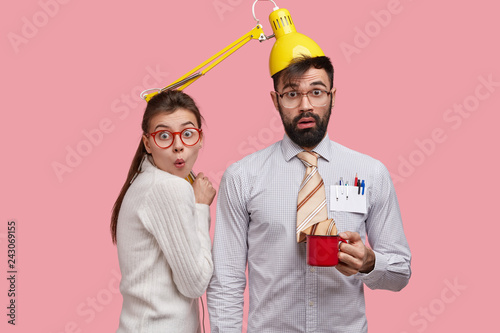 Photo of funny students collaborate together, have fun during coffee break, surprised expressions. Handsome male teacher in formal clothes has lamp on head, gives private lesson to female trainee photo