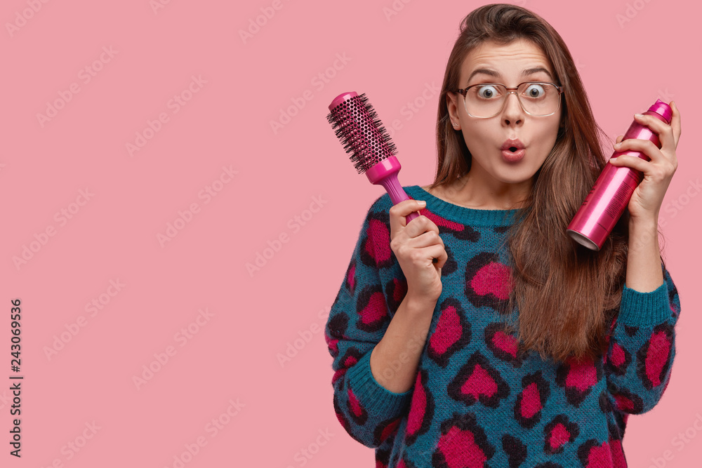 Astonished young lady has mess on head, prepares for special occasion, makes coiffure, surprised to look at herself in mirror, carries comb and spray, isolated over pink wall with blank space Astonished young lady has mess on head, prepares for special occasion, makes coiffure, surprised to look at herself in mirror, carries comb and spray, isolated over pink wall with blank space