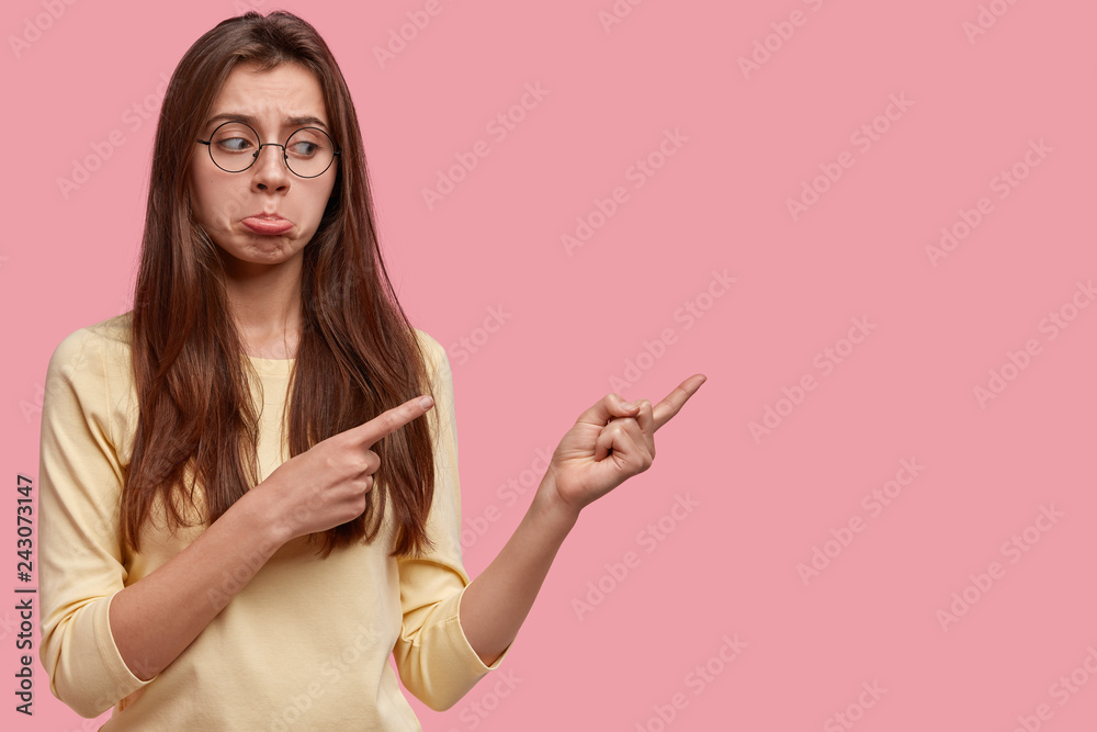 Depressed young woman purses lower lip, indicates at something on pink studio wall, has displeased facial expression, looks with dissatisfaction and frustration, doesnt like item. Look aside Depressed young woman purses lower lip, indicates at something on pink studio wall, has displeased facial expression, looks with dissatisfaction and frustration, doesnt like item. Look aside