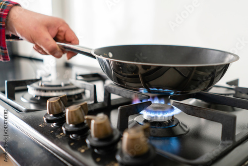 A man cooks in a frying pan, puts it on the stove. Modern gas burner and hob on a kitchen range. Dark black color and wooden Small kitchen in a modern apartment