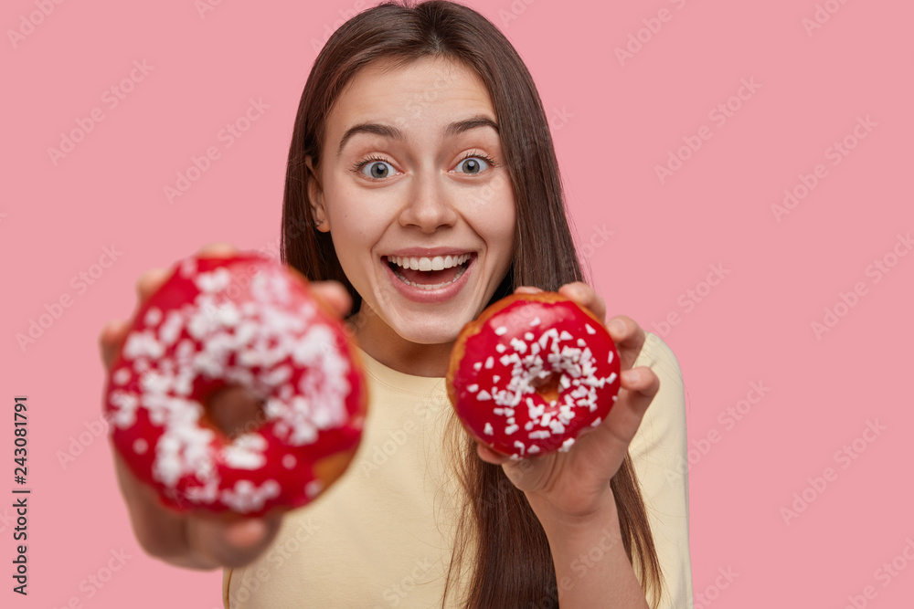 Glad happy young woman has broad smile, being in high spirit, carries tasty dessert, focus on douchnuts, wears casual clothes, isolated over pink background. People, eating, dieting concept. Glad happy young woman has broad smile, being in high spirit, carries tasty dessert, focus on douchnuts, wears casual clothes, isolated over pink background. People, eating, dieting concept.