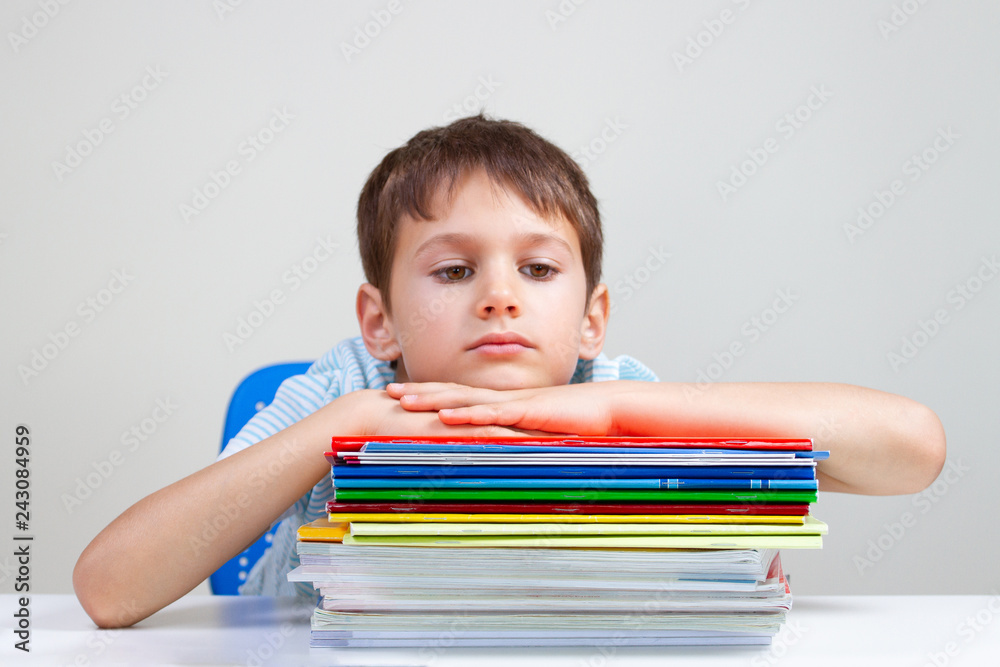 Upset schoolboy sitting at desk with pile of school books and notebooks Upset schoolboy sitting at desk with pile of school books and notebooks