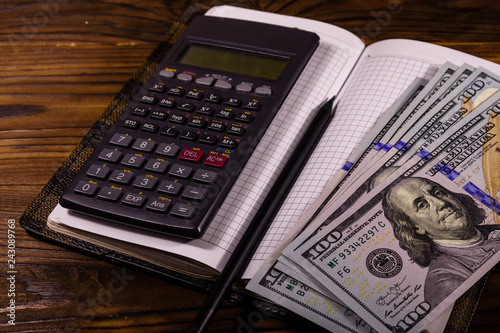 Opened notepad, one hundred dollar banknotes, scientific calculator and pencil on wooden table