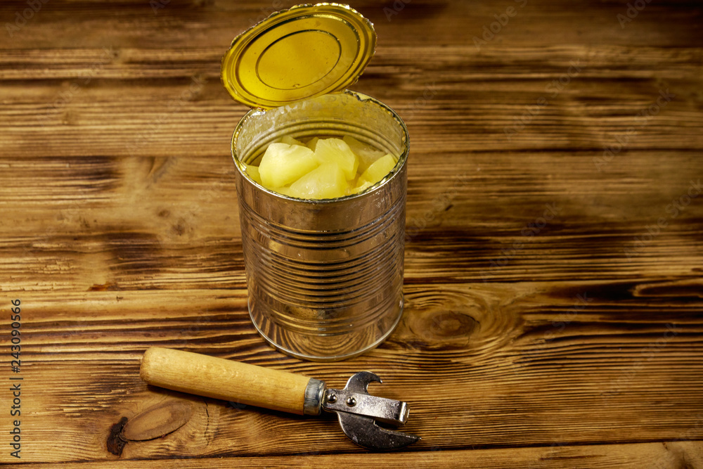 Opened tin can of pineapple pieces and can opener on wooden table