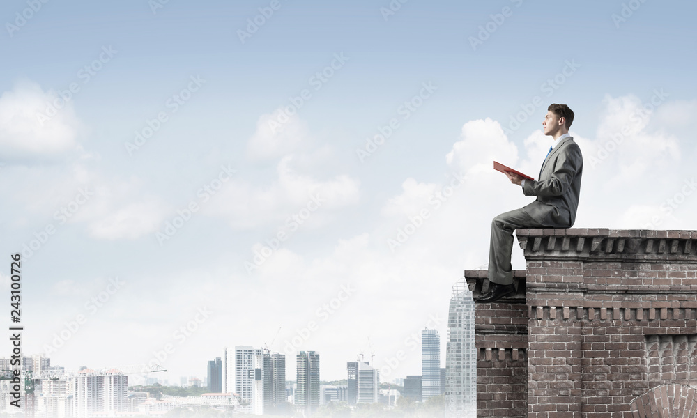 Man on roof edge reading book and cityscape at background Man on roof edge reading book and cityscape at background