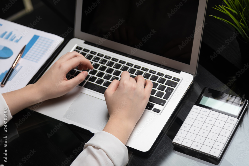 Close-up of hands typing on laptop with blank black screen next to charts and calculator Close-up of hands typing on laptop with blank black screen next to charts and calculator