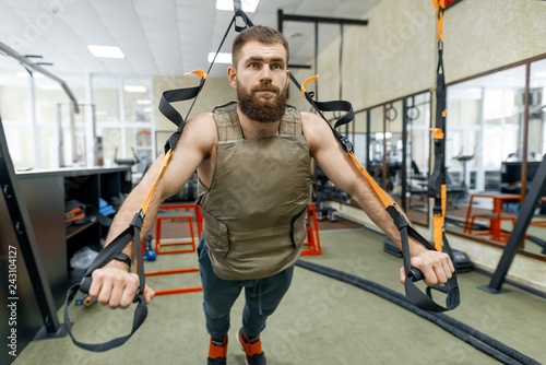 Muscular bearded man dressed in military weighted armored vest doing exercises using straps systems in the gym. Sport, training, bodybuilding and healthy lifestyle concept.