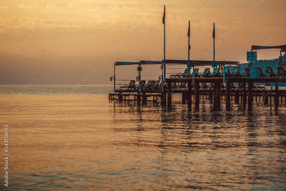 Stunning view of the pier with closed umbrellas and deck chairs on it at beautiful sunset reflecting in the calm sea