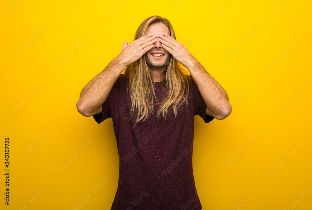 Blond man with long hair over yellow wall covering eyes by hands. Surprised to see what is ahead Blond man with long hair over yellow wall covering eyes by hands. Surprised to see what is ahead