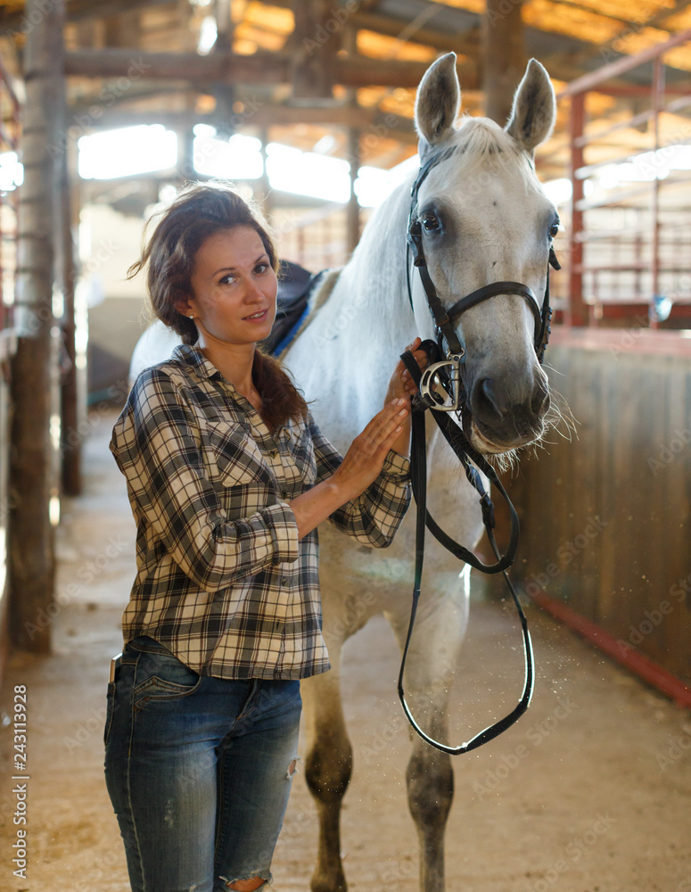 Obraz premium Woman farmer standing with white horse at stabling indoor