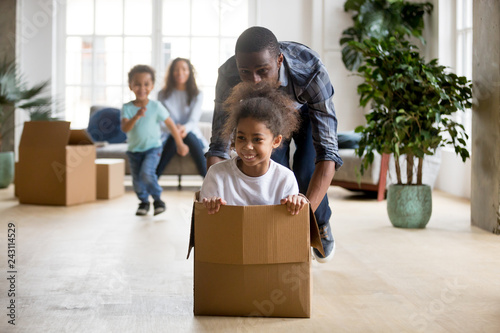 African dad playing with mixed race daughter riding in box on moving day concept, little kids enjoy game with father in living room, happy black family and children having fun in new home, relocation