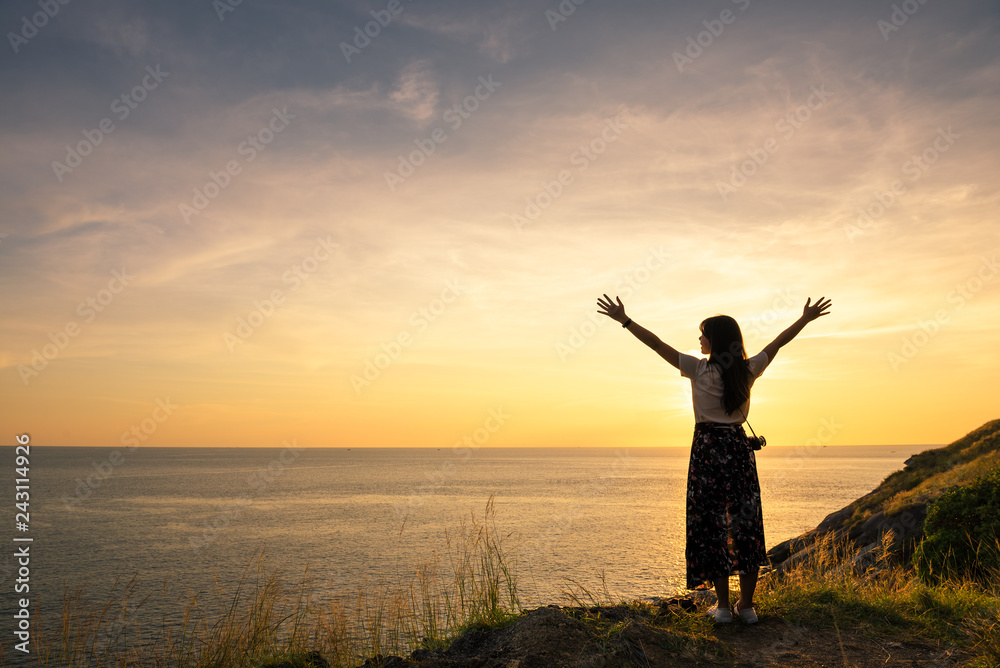 Happy young woman enjoying freedom with open hands and sitting on stone and sunset on the sea sky sunset background