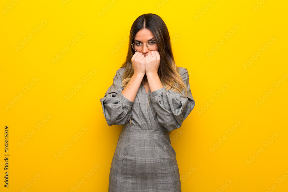Young woman with glasses over yellow wall is a little bit nervous and scared putting hands to mouth Young woman with glasses over yellow wall is a little bit nervous and scared putting hands to mouth