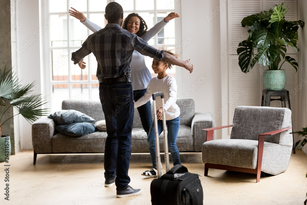 Happy loving black family wife and kid daughter excited to meet hurry to hug dad with suitcase coming returning home arriving from long business trip, welcome back african father, reunion concept