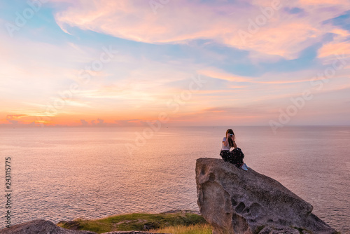 young woman sit on stone and use mobile phone takes a photo with beautiful amazing sea sunset of orange red colors