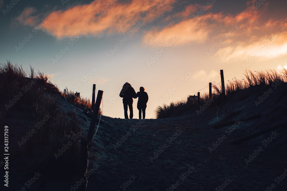Love couple take a winter vacation walk. Silhouette of two person on beach sand dunes with sunset colorful sky tones. German Baltic Sea coastline at Fischland-Darss-Zingst in Mecklenburg