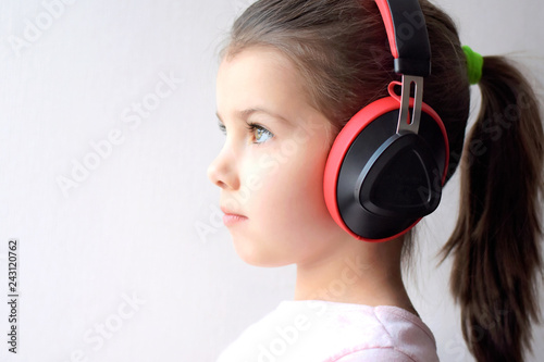 Portrait of caucasian white pretty little girl which listening to music with red headphones. Beautiful brunette green eyed girl with pony tail hair listen to music with big earphones.