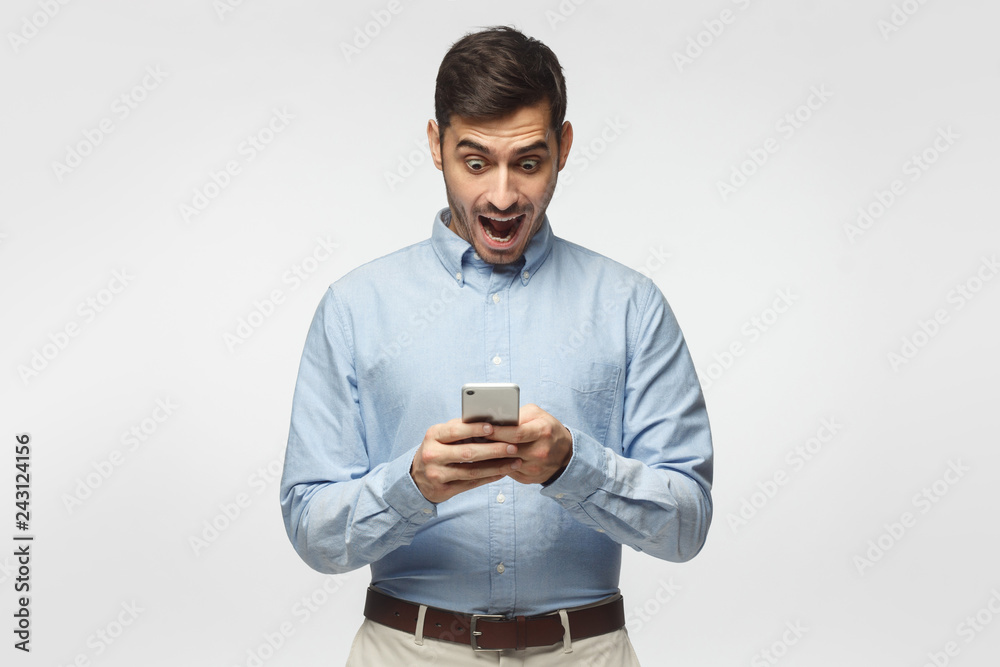 Young business man looking at his phone with surprise expression, isolated on gray background Young business man looking at his phone with surprise expression, isolated on gray background