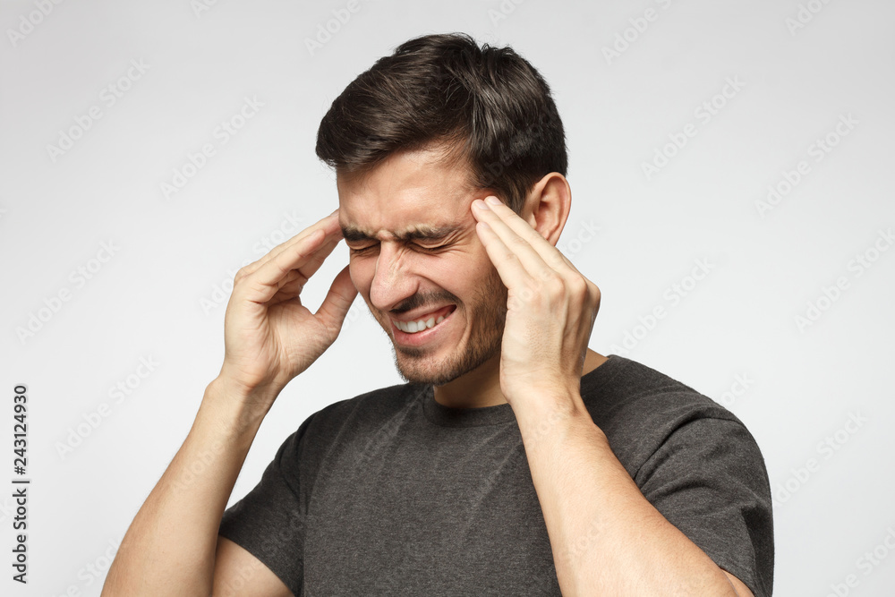 Close-up of young man isolated on gray background in casual gray t-shirt, touching temples with fingers as if suffering from severe migraine Close-up of young man isolated on gray background in casual gray t-shirt, touching temples with fingers as if suffering from severe migraine