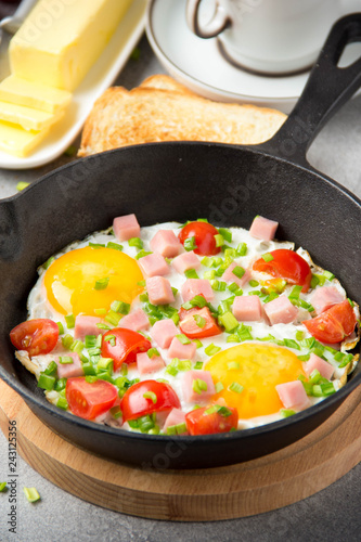 Fried eggs in black pan with ham, cherry tomatoes and spring green onions. Delicious Breakfast with toast and butter. The classic English lunch