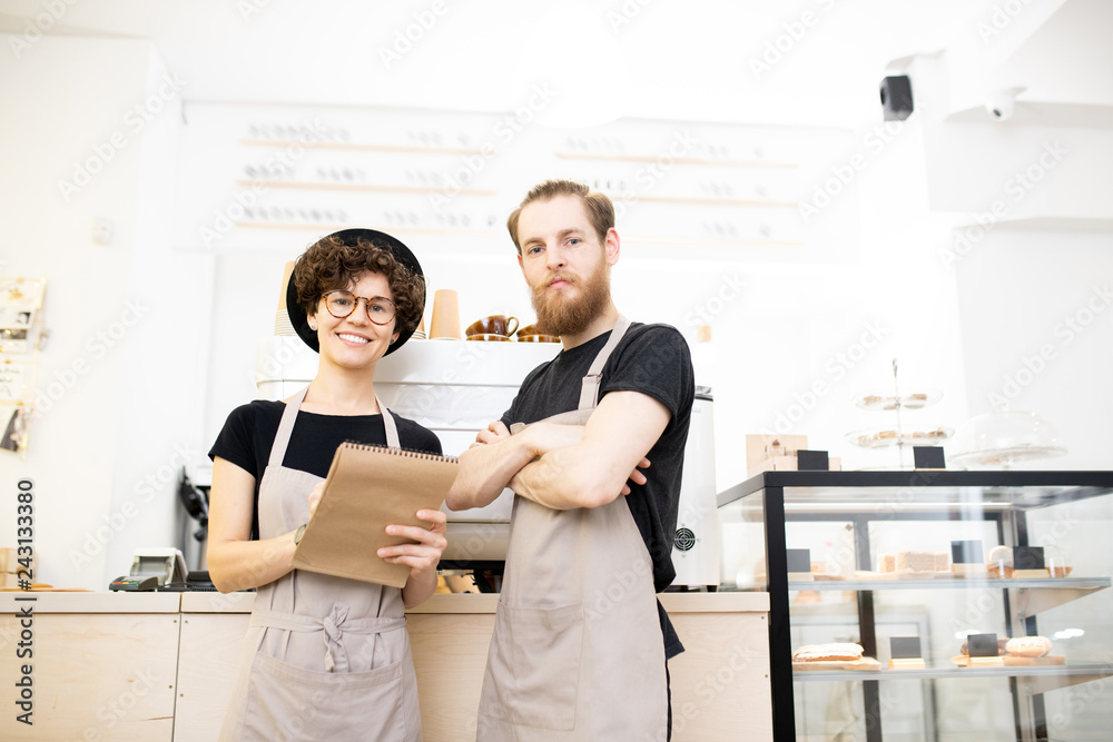 Fototapeta premium Content confident team of coffee shop: smiling young man and woman in aprons standing at counter and looking at camera while analyzing notes in sketchpad