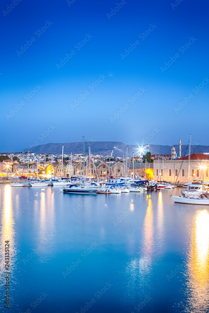Travel Concepts. Picturesque Image of Old Venetian Harbour of Chania with line of Fisihing Boats and Yachts in the Foregound At Blue Hour in Crete.