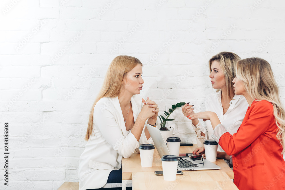 Group of young women co-workers discussing creative project during work process, sitting at office with laptop and take-away coffee. Blonde business woman explains main ideas to her colleagues. Group of young women co-workers discussing creative project during work process, sitting at office with laptop and take-away coffee. Blonde business woman explains main ideas to her colleagues.
