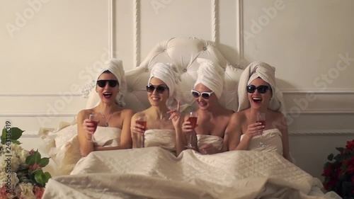 Four girlfriends are in the same bed with towels on their heads. girls laugh and drink champagne