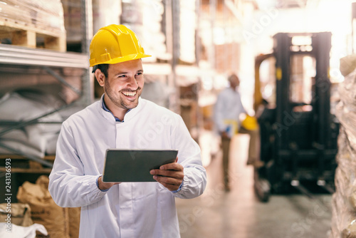 Close up of young smiling bearded man with protective helmet on head using tablet while standing in warehouse.