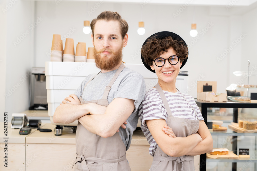 Positive confident hipster couple of baristas in aprons standing against espresso machine and looking at camera Positive confident hipster couple of baristas in aprons standing against espresso machine and looking at camera