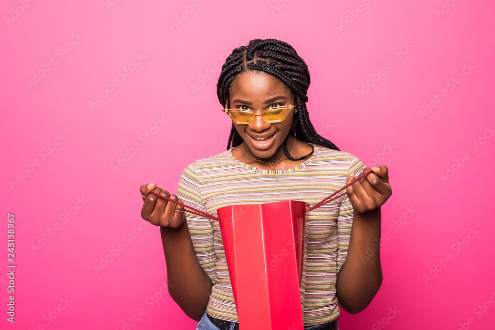 Portrait of beautiful african american girl looking into shopping bag with surprised expression on face isolated on pink background