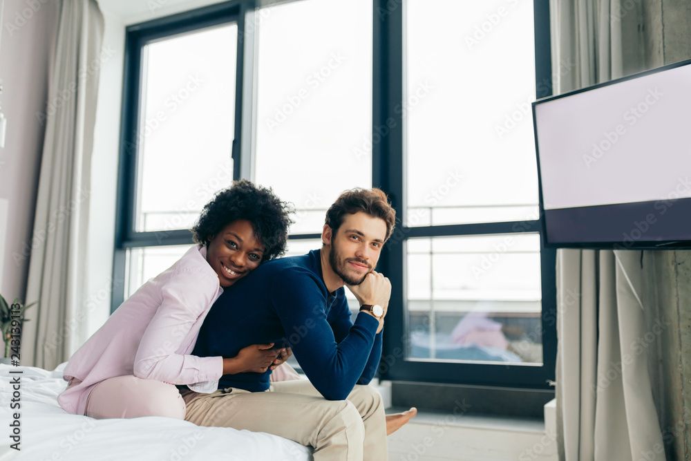 Happy African american girl embracing her handsome caucasian boyfriend while sitting on bed in bedroom with modern grey colored interior and big windows.