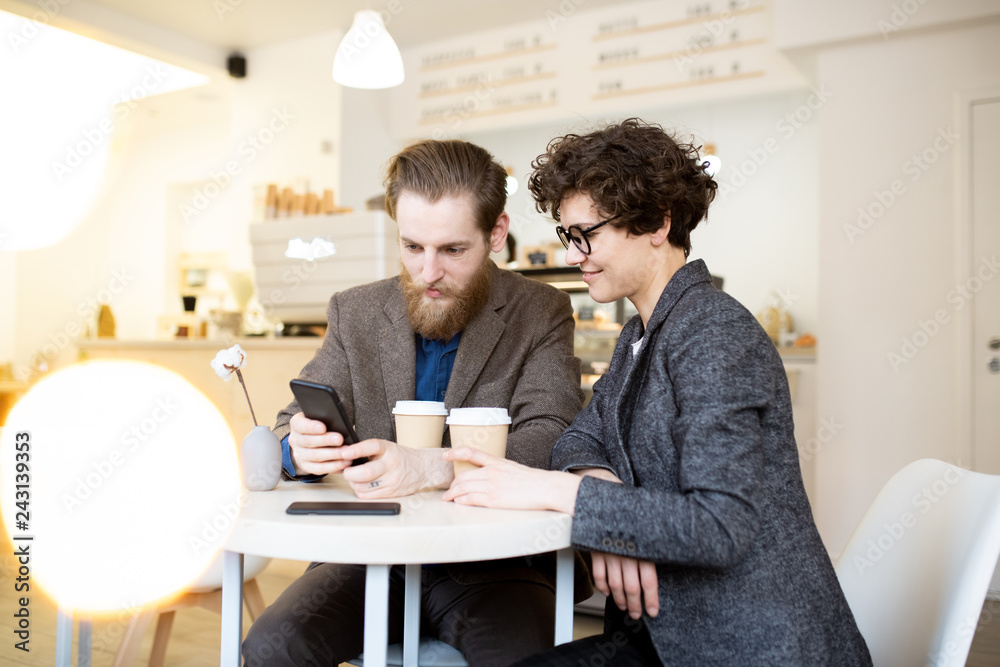 Positive young couple in gray jackets sitting at small table in cozy cafe and viewing online source on phone while discussing social media project
