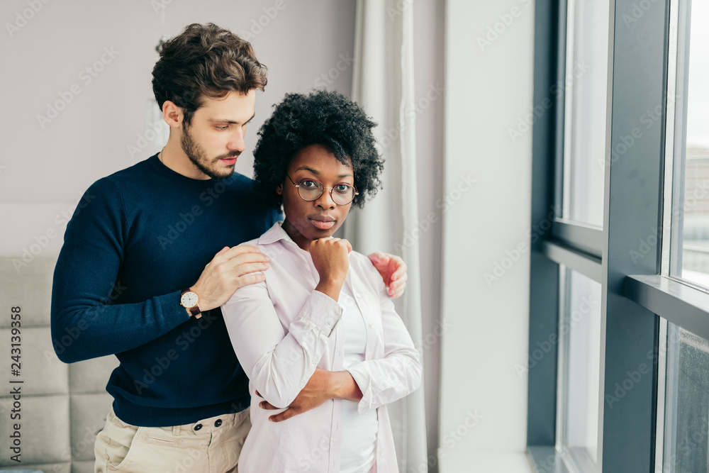 Interracial merried couple embrace affectionately at home interior, sitting near large window, feel support and emotional resource from each other, have close relationship.