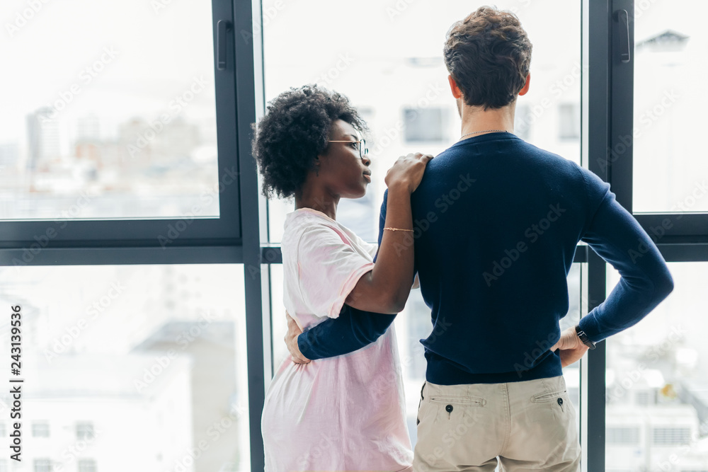 Backside view of young mixed race lovers staying in hotel, Hispanic man hugging African Woman while resting in modern apartment with big window interior Backside view of young mixed race lovers staying in hotel, Hispanic man hugging African Woman while resting in modern apartment with big window interior