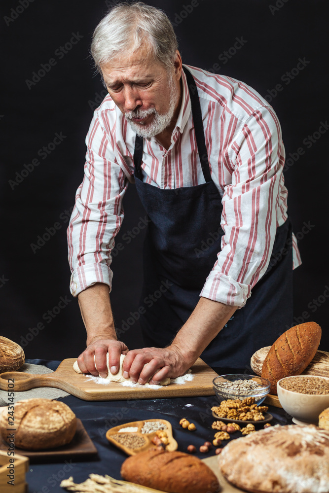 Senior Man, Baker Chef cooking bread in the studio over black background. Grandpa kneading sour dough for baking rustic organic bread. Natural light, moody still life. Senior Man, Baker Chef cooking bread in the studio over black background. Grandpa kneading sour dough for baking rustic organic bread. Natural light, moody still life.