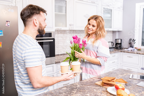 Waist up portrait of happy young couple at home in kitchen, focus on smiling woman holding bouquet of pink roses