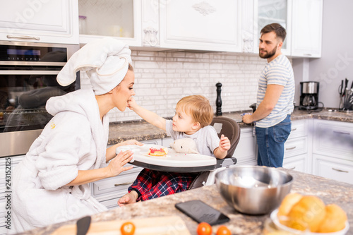 Portrait of happy family in kitchen enjoying breakfast at home, focus on woman feeding baby in foreground