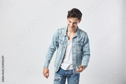 Smiling young dark-haired guy in a white t-shirt, jeans and a denim jacket stands on the white background  in the studio © Leika production