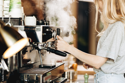 A young pretty thin blonde,dressed in casual outfit,is cooking coffee in a popular coffee shop.
