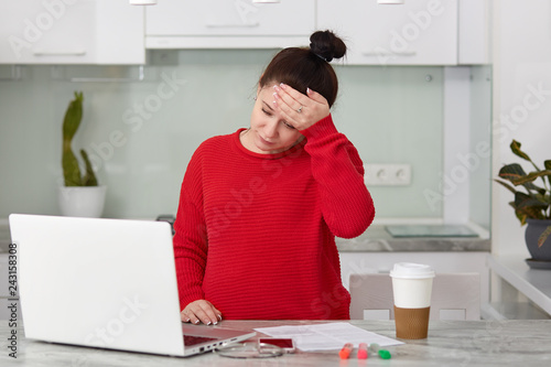 Photo of pregnant Caucasian woman has headache, keeps hand of forehead, wears casual red sweater, works on laptop computer, develops her personal blog, uses internet, sits at kitchen interior