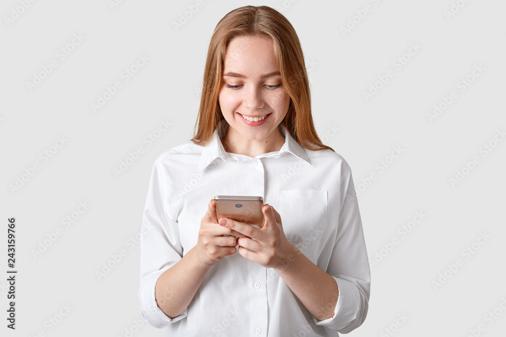 Positive young girl with long strainght hair, holds cell phone, recieves text message, dressed in elegant white shirt in one tone with background. Happy youngster surfes social networks online Positive young girl with long strainght hair, holds cell phone, recieves text message, dressed in elegant white shirt in one tone with background. Happy youngster surfes social networks online